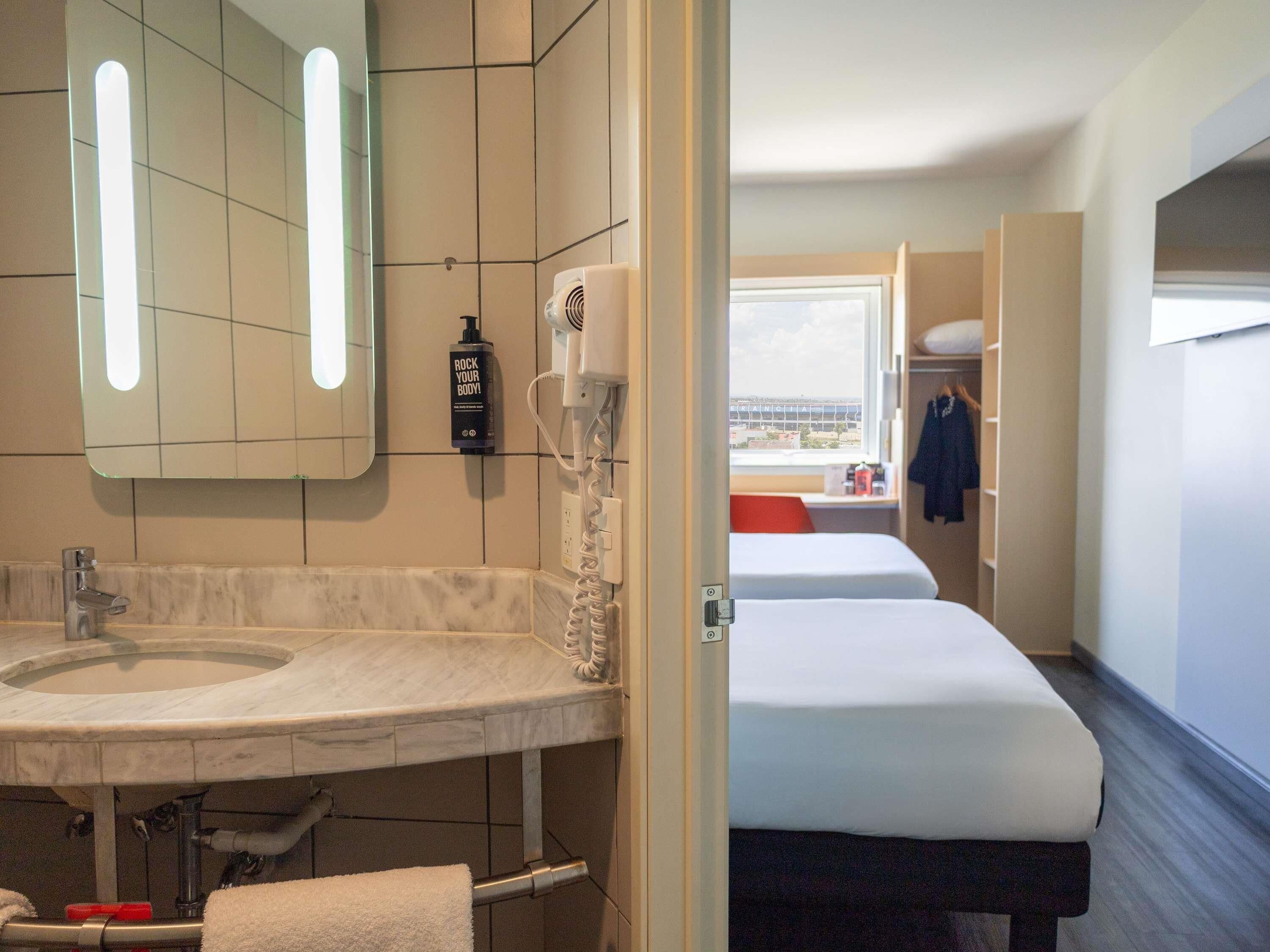 Baño de hotel con lavamanos, espejo y secador de pelo. Se ve una ventana con vistas a edificios.