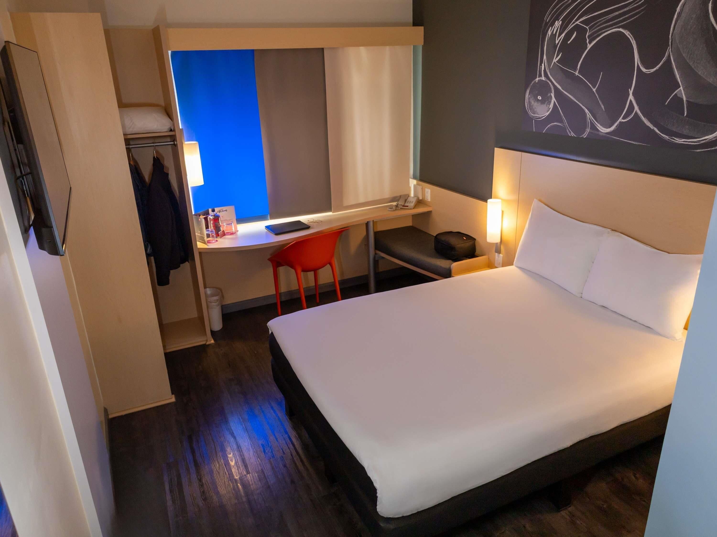 A hotel room with a bed, a desk with a chair, a small table, and a wall-mounted artwork.