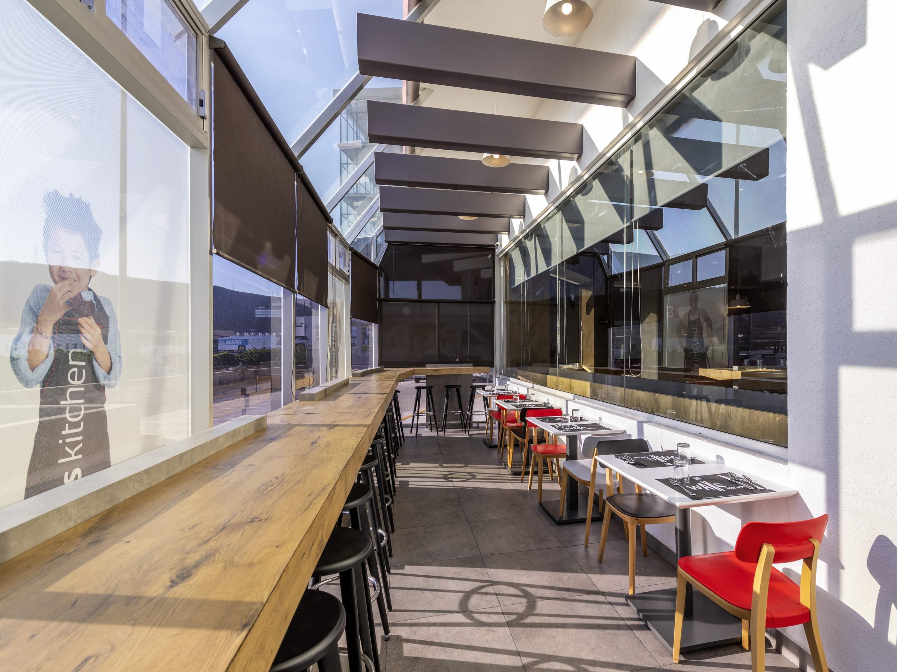 Un restaurant moderne doté d’un long comptoir en bois, de chaises rouges et en bois, et de grandes fenêtres offrant une vue sur la ville.