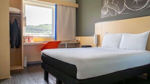 A hotel room with a bed, a desk, a chair, a window with curtains, a coat rack, and a wall art piece. - ibis Queretaro (Querétaro)