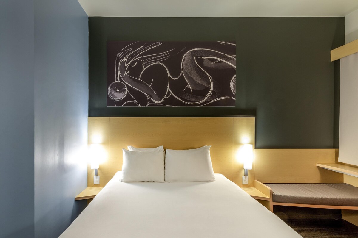 A hotel room with a large bed, two bedside lamps, a wooden headboard, and a wall art piece above the bed.