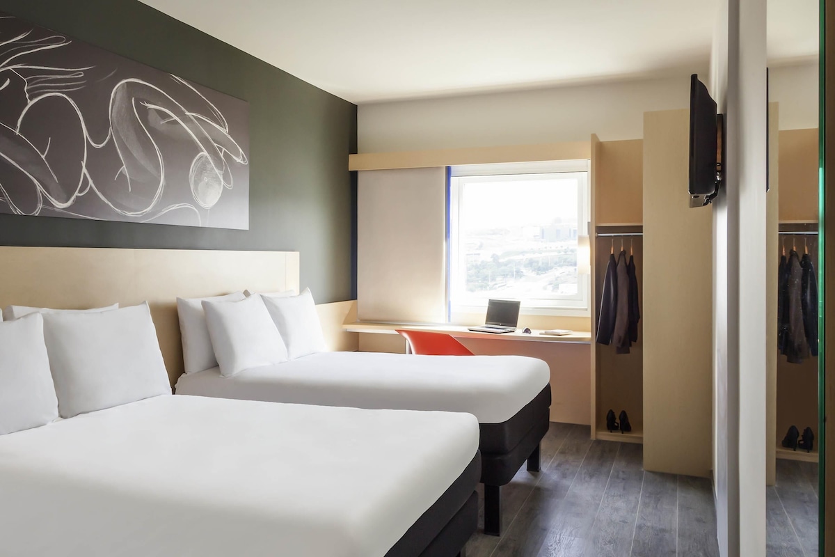 Habitación de hotel con dos camas, un escritorio con una computadora portátil, un perchero y una ventana con vista a la ciudad.