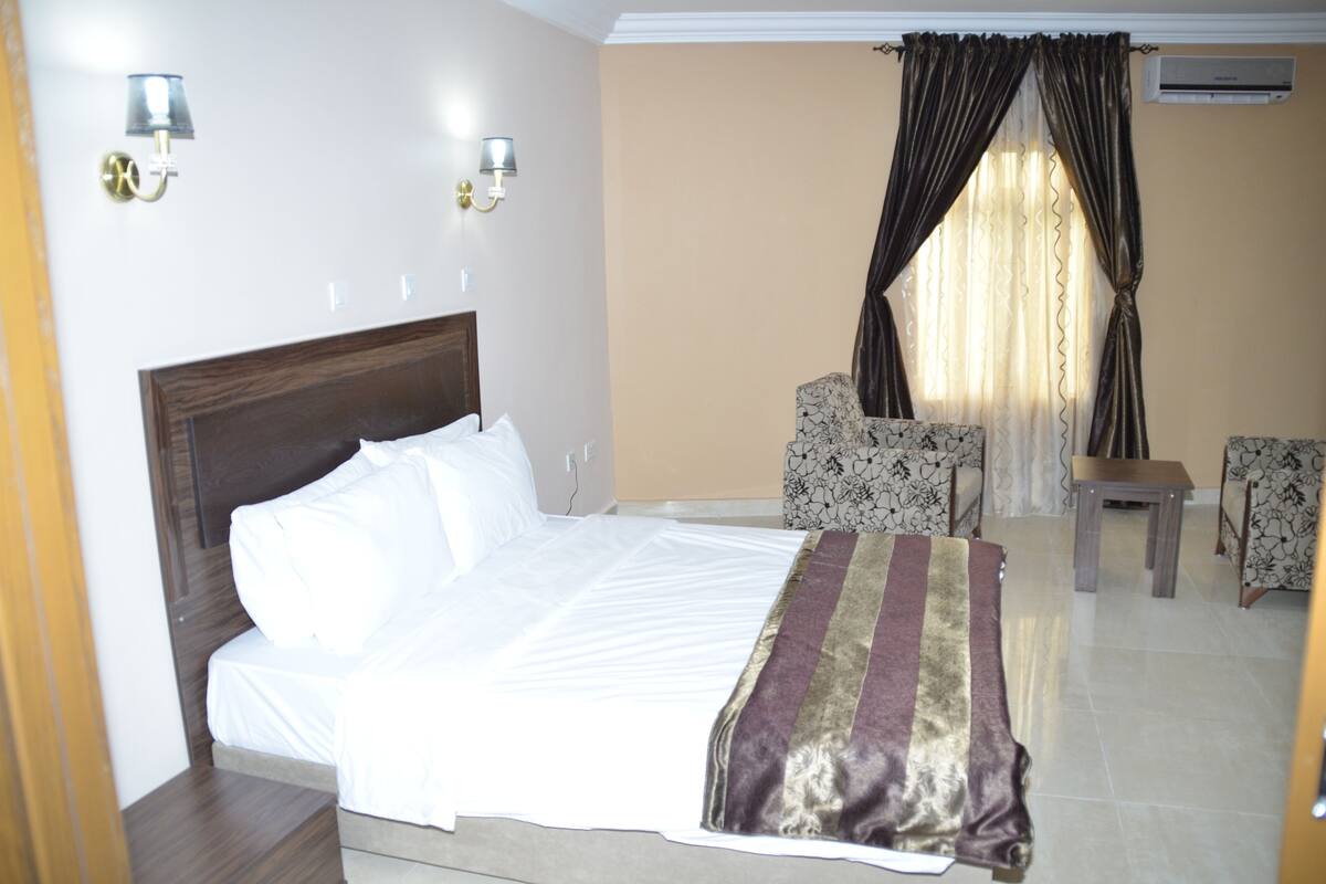 A hotel room with a bed, two chairs, a small table, and a window with curtains.