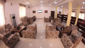 A spacious room with a central table surrounded by floral-patterned sofas, a flat-screen TV mounted on the wall, and a large pillar in the background. - Residency Hotel Leophine House (Ogbunike)