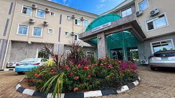 A hotel entrance with a green canopy, air conditioning units, and a flower bed in the foreground.
