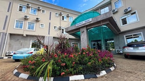 L’entrée d’un hôtel, avec un auvent vert, des unités de climatisation et un parterre de fleurs au premier plan.