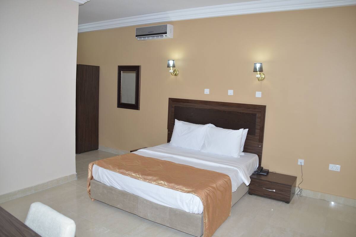A bedroom with a bed, bedside table, nightstand, wall-mounted lamp, and an air conditioning unit.