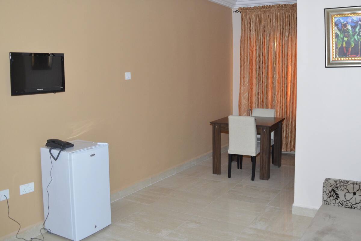 A living room with a flat-screen TV mounted on the wall, a small white refrigerator, a dining table with two chairs, and a framed picture on the wall.