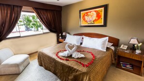 Quarto de hotel com cama, mesa de cabeceira, abajur, quadro na parede e janela com cortinas.
