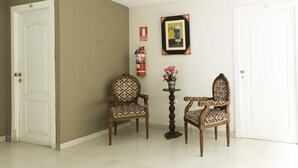Um corredor com duas cadeiras de madeira, uma pequena mesa com uma planta em vaso e um quadro na parede.