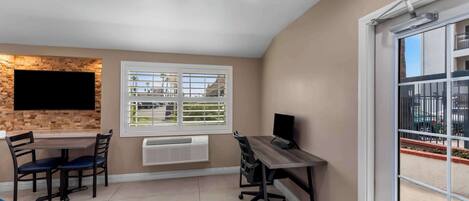 Una habitación con una mesa de comedor, sillas, un televisor fijado a una pared de ladrillo, una ventana con persianas y un balcón con barandilla.