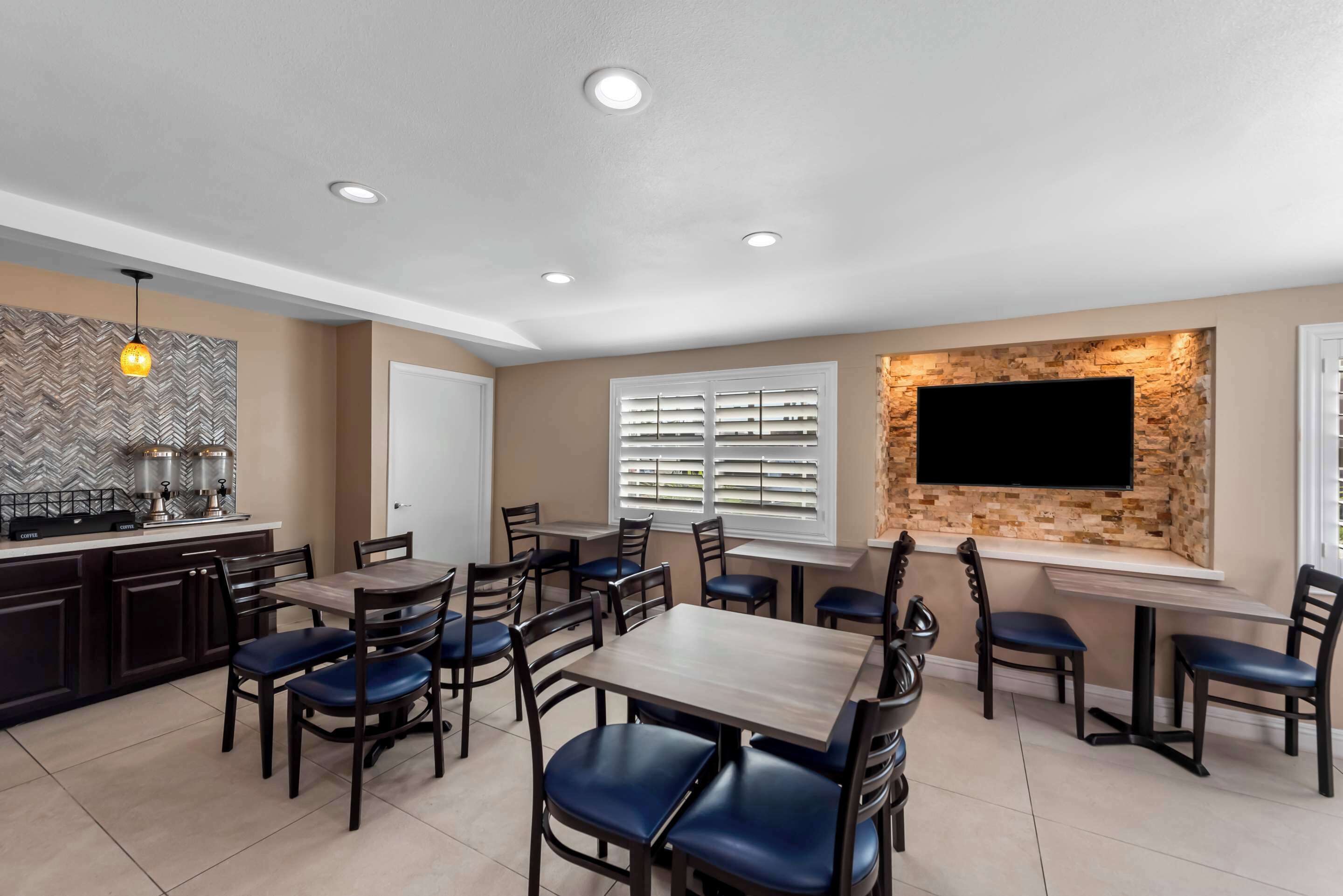 A dining area with multiple tables and chairs, a kitchen area with dark cabinets, and a mounted flat-screen TV on the wall.