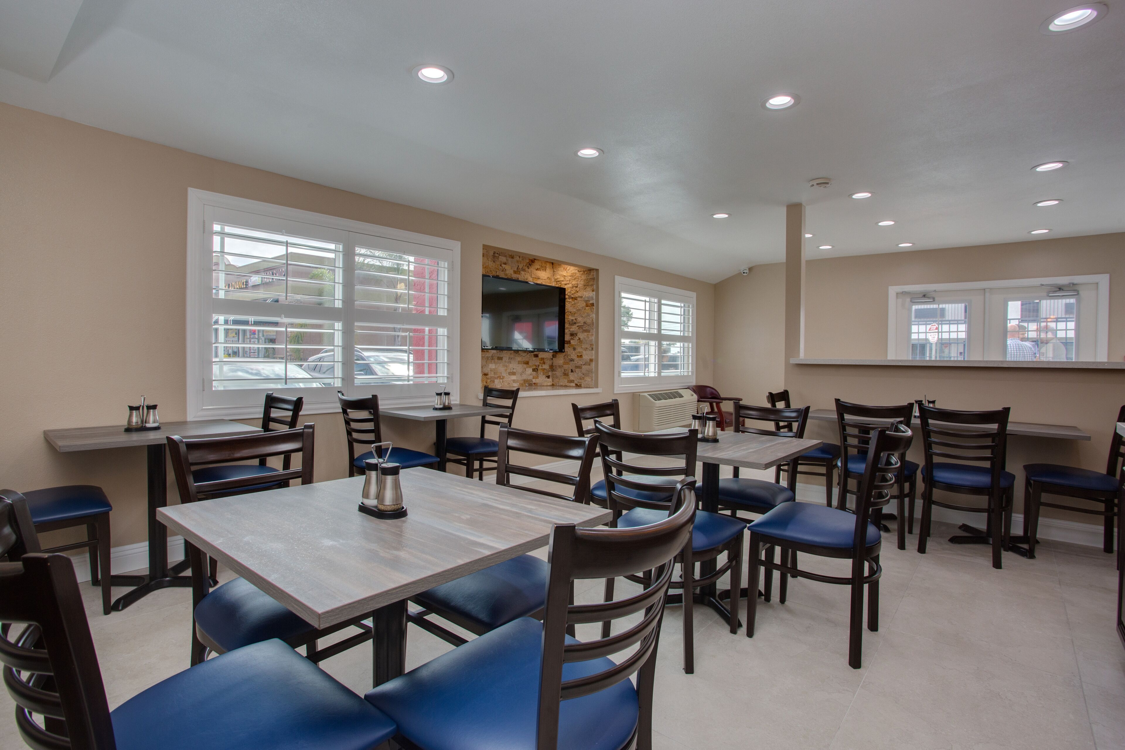 A restaurant with multiple tables and chairs, a flat-screen TV mounted on a stone wall, and large windows with blinds.