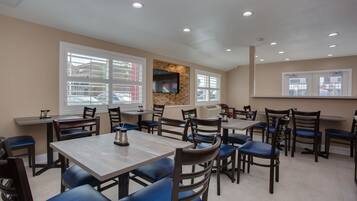 A restaurant with multiple tables and chairs, a flat-screen TV mounted on a stone wall, and large windows with blinds.