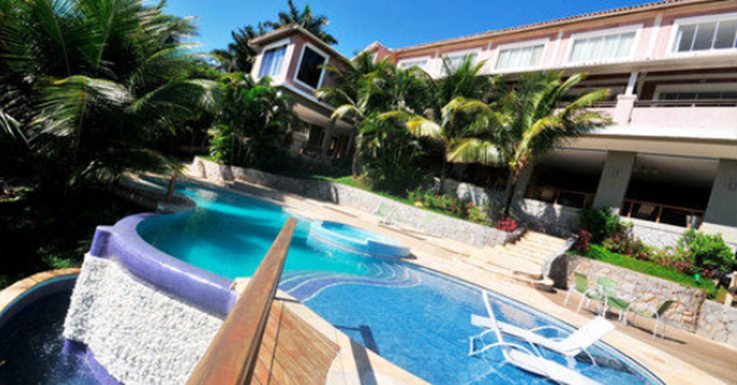 A swimming pool with lounge chairs and a hot tub, surrounded by palm trees and a building with a red roof.