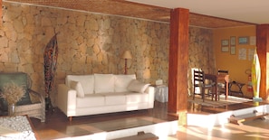 Una habitación con pared de piedra, un sofá blanco, un sillón de mimbre, una mesa de madera y una lámpara.