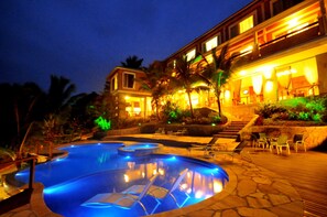 Una piscina con sillas de descanso y un edificio con ventanas iluminadas y zona de estar al aire libre.
