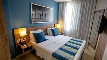 Habitación de hotel con una cama grande, dos mesitas de noche con lámparas, un televisor montado y un cuadro en la pared.