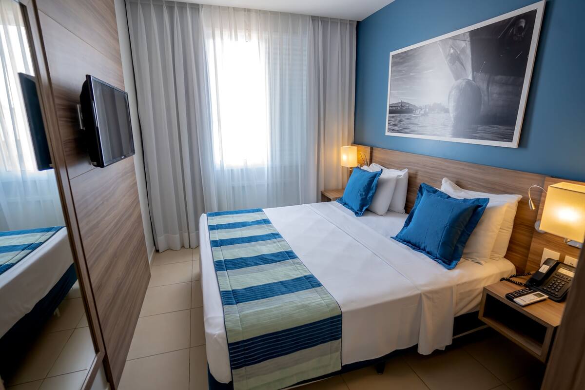 A hotel room with a bed, a TV mounted on the wall, a bedside table with a lamp, and a painting on the wall.