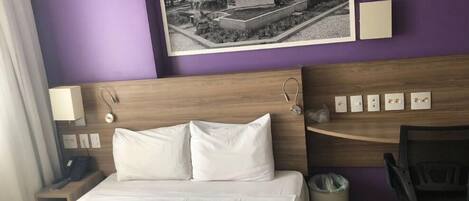 Habitación de hotel con cama, escritorio, silla, teléfono y cubo de basura.