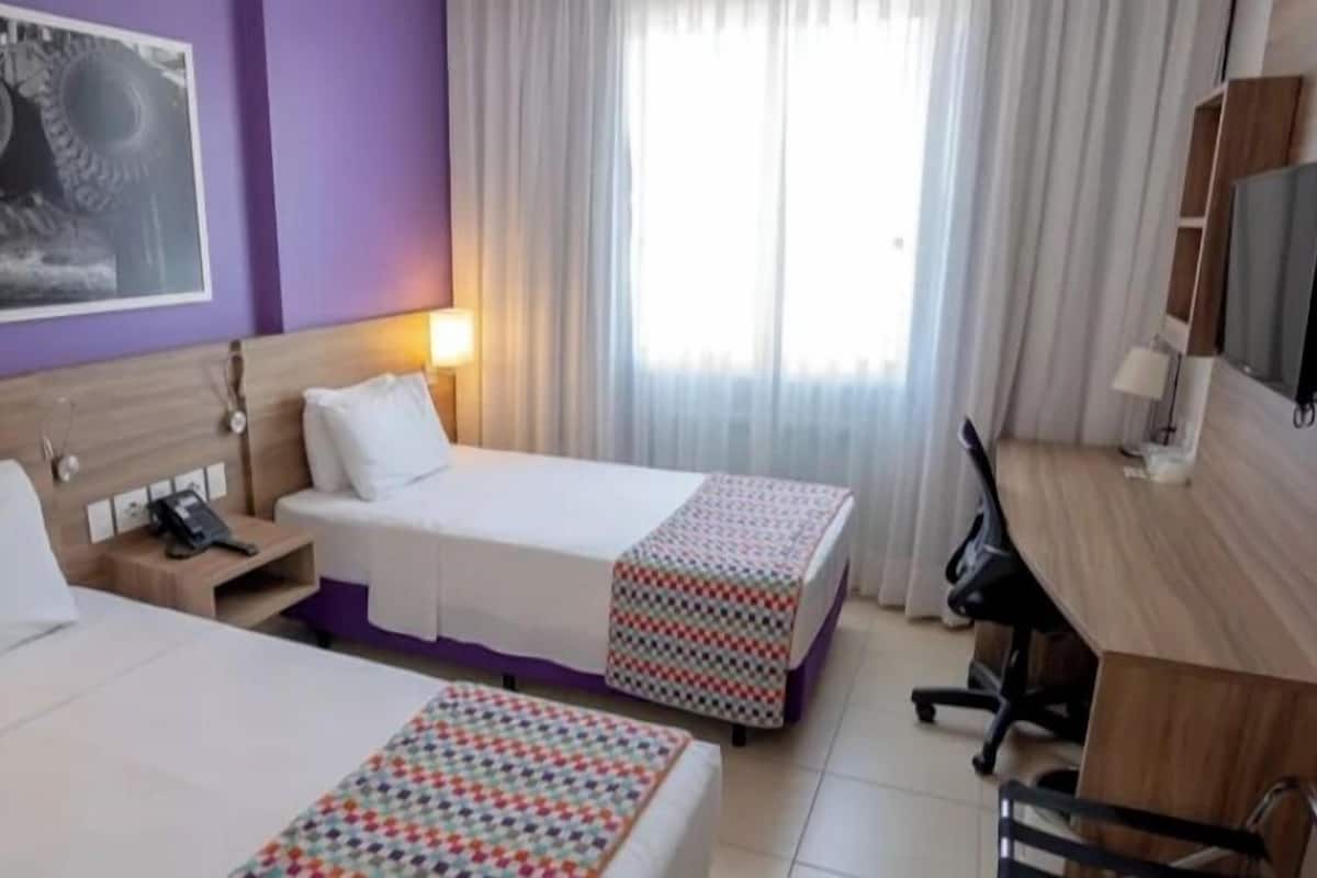 A hotel room with two beds, a desk, a chair, a TV, and a window with curtains.