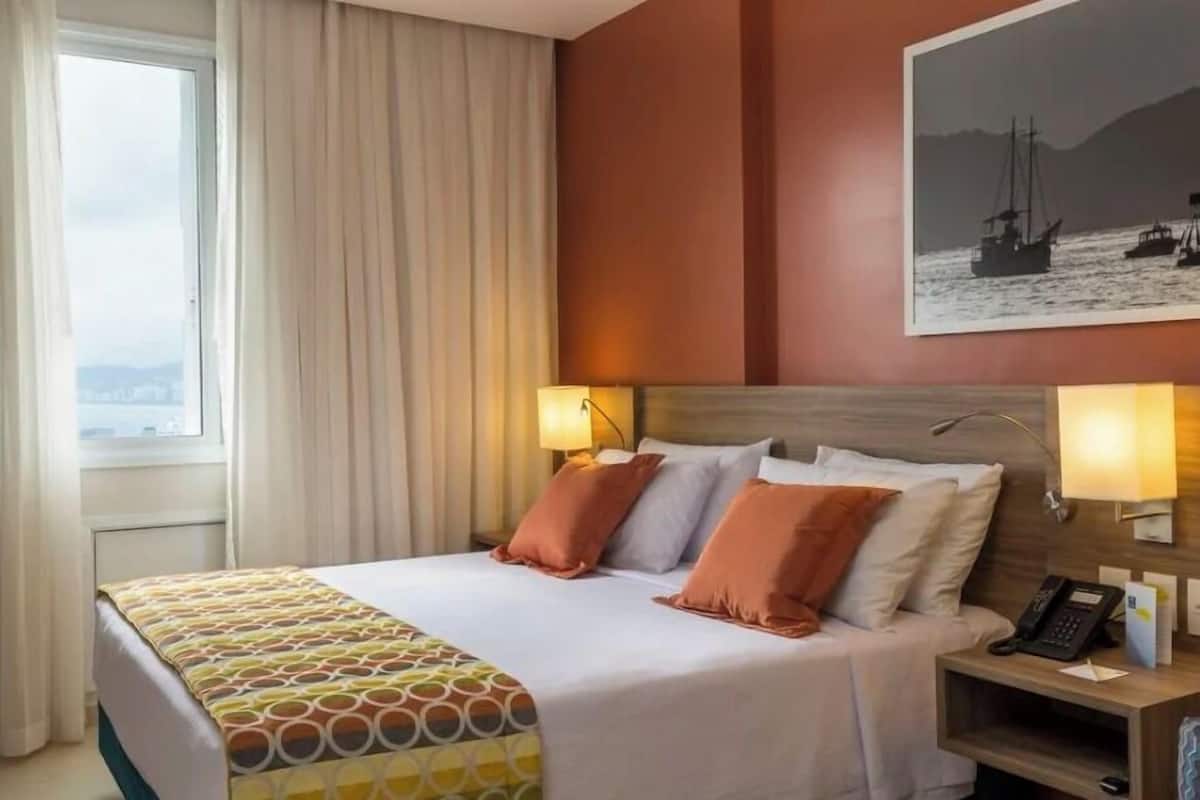 A hotel room with a bed, two bedside lamps, a nightstand with a phone, and a window with curtains.