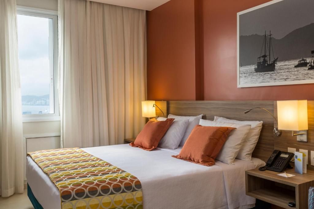 A hotel room with a bed, two bedside lamps, a nightstand with a phone, and a window with curtains.