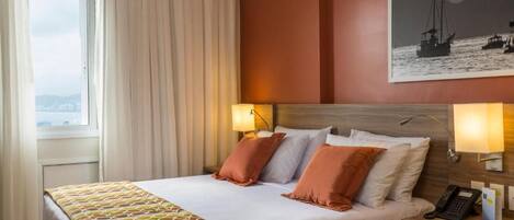 Habitación de hotel con cama, dos lámparas de noche, mesita de noche con teléfono y ventana con cortinas.