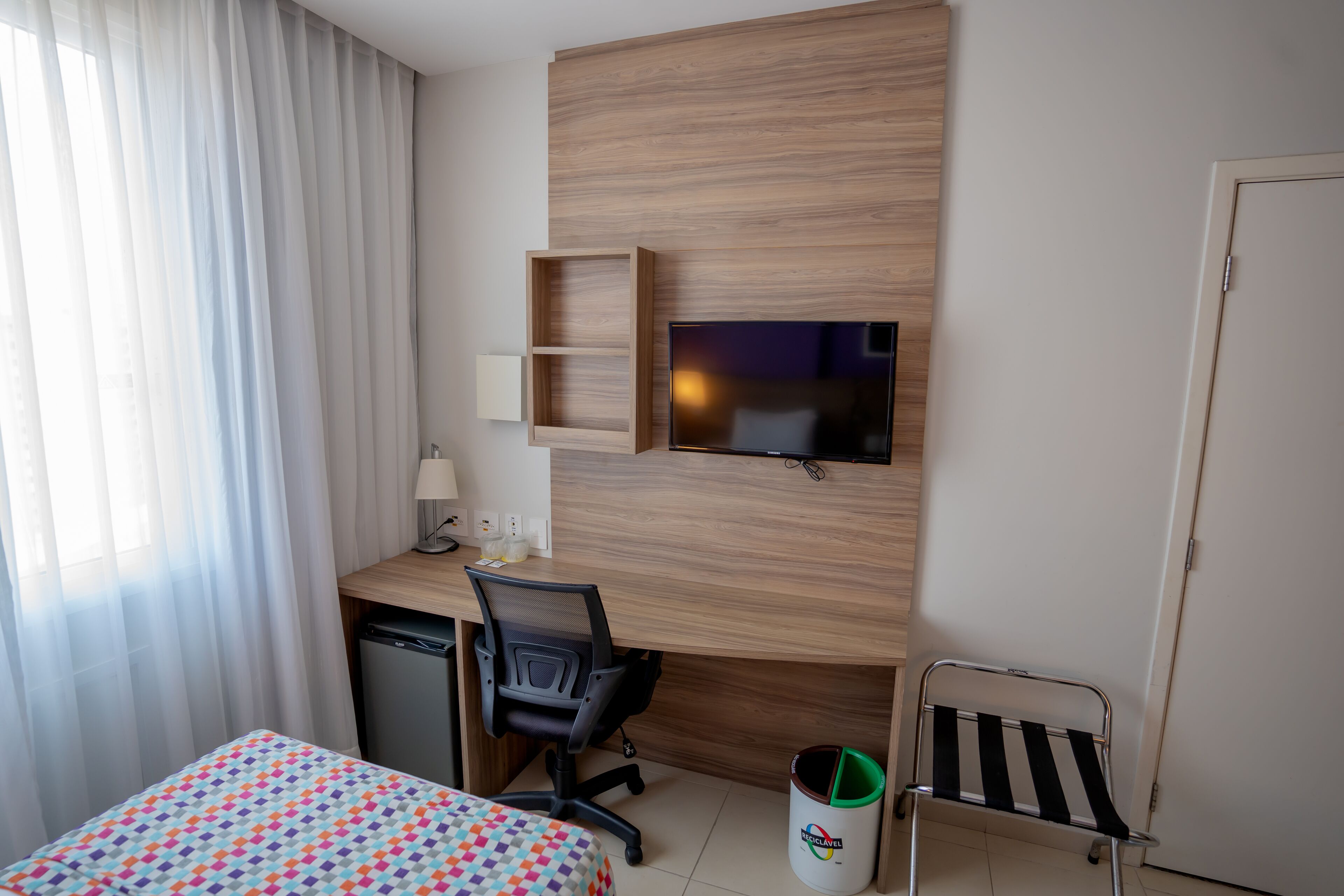Een hotelkamer met een bed, bureau, stoel, televisie en een kleine prullenbak.