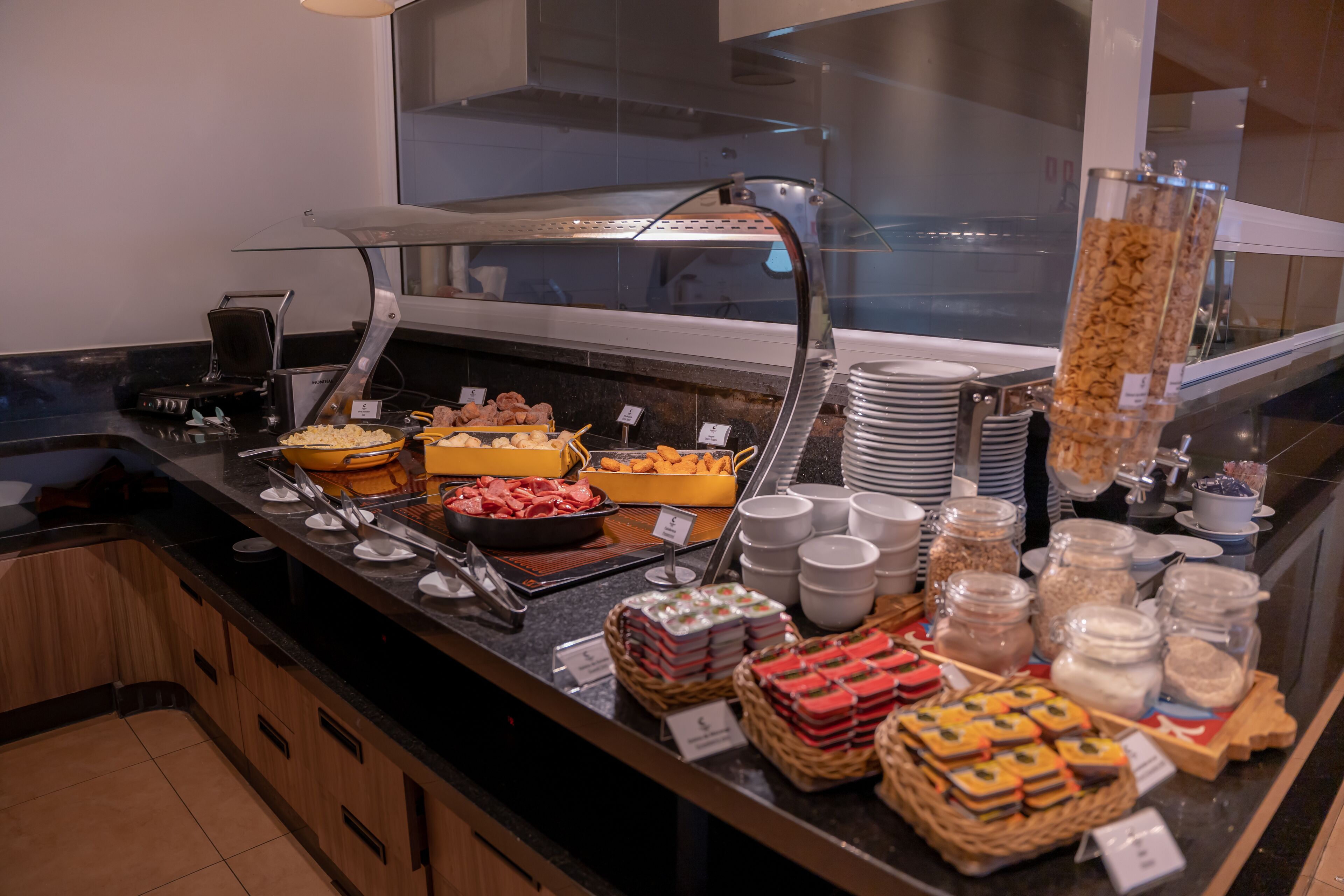 Un bufé con diversos alimentos, incluyendo carnes, quesos y pasteles, dispuesto en una barra con utensilios para servir y platos.