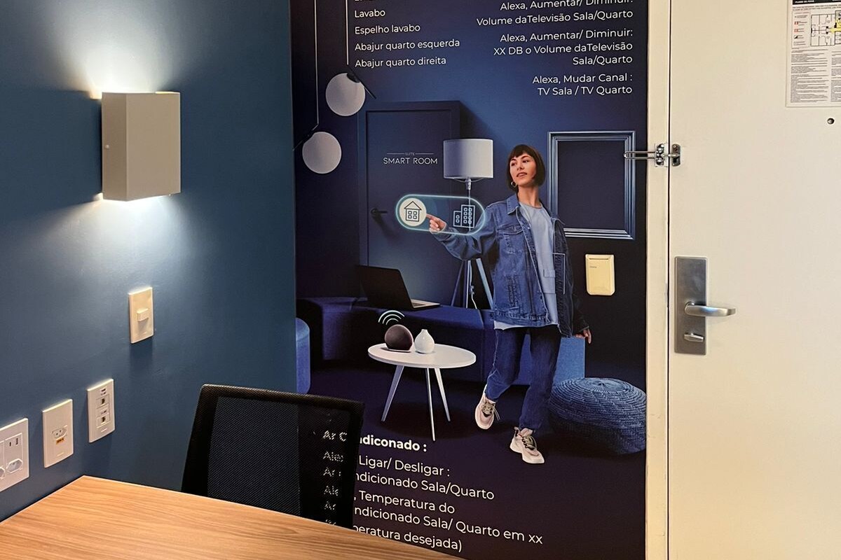 A modern room with a blue wall, a wooden table, and a white door. There is a wall-mounted light fixture and a poster with a person demonstrating a smart home device.