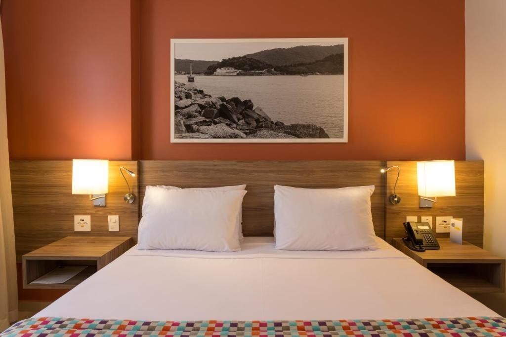 Una habitación de hotel con una cama, dos lámparas de noche, una mesita de noche con un teléfono y un cuadro en la pared.
