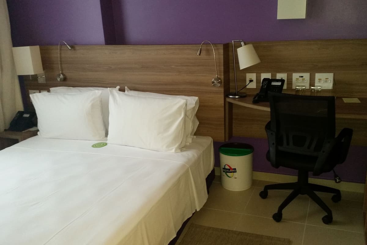 A hotel room with a bed, a desk, a chair, a trash bin, and a wall-mounted phone.