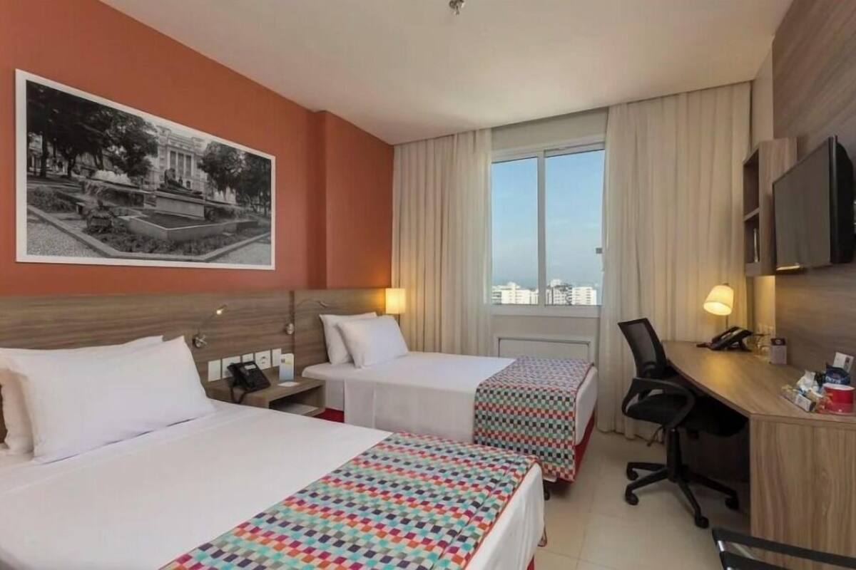 A hotel room with two beds, a desk, a chair, a TV, and a large window with curtains.