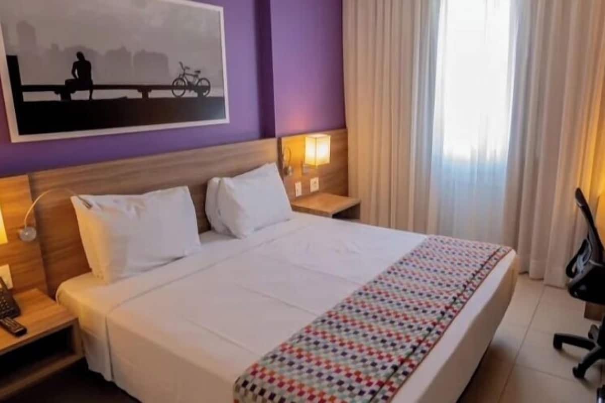A hotel room with a large bed, two bedside tables with lamps, a desk with a chair, and a window with curtains.