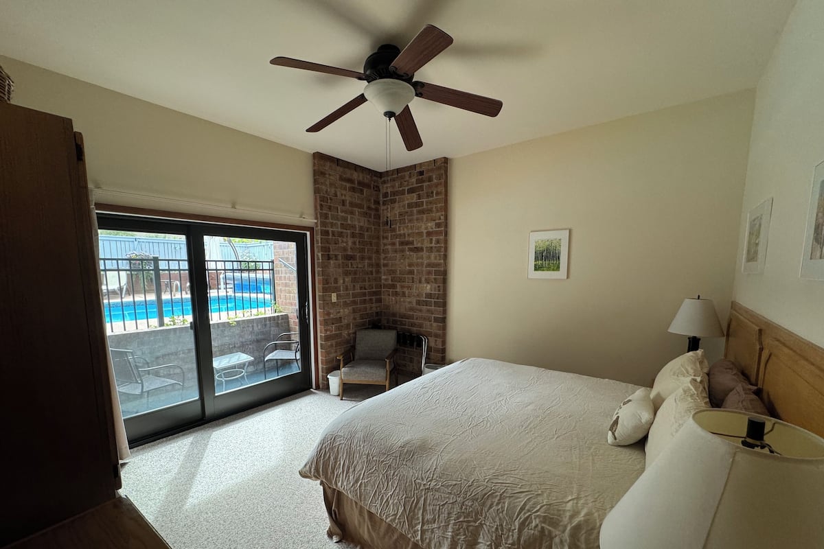 Une chambre d’hôtel avec un lit, une chaise, un ventilateur de plafond, une fenêtre donnant sur une piscine et un mur en briques.