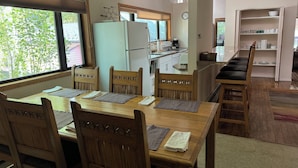 Un comedor con una mesa y sillas de madera, una cocina al fondo y vista a árboles fuera.