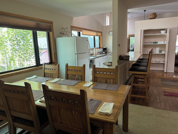 A dining area with a wooden table and chairs, a kitchen in the background, and a view of trees outside. - Shadow Mountain Lodge (Aspen)
