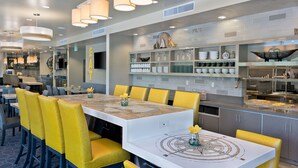 A modern restaurant interior with a central dining area featuring a white table with a geometric table runner, yellow chairs, and pendant lighting. - Oxford Suites Bellingham (Bellingham)