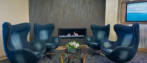 Une pièce avec une cheminée, deux fauteuils bleus, une petite table sur laquelle est posé un vase de fleurs et un téléviseur fixé au mur.