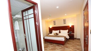A hotel room with a wooden bed, a bedside table, a nightstand, a wall-mounted lamp, and a shower area with a glass door. - Petra Venus Hotel (Wadi Musa)