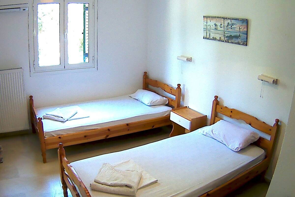 A room with two wooden beds, each with a pillow and a folded towel. There is a window with blinds, a radiator, and a small painting on the wall.