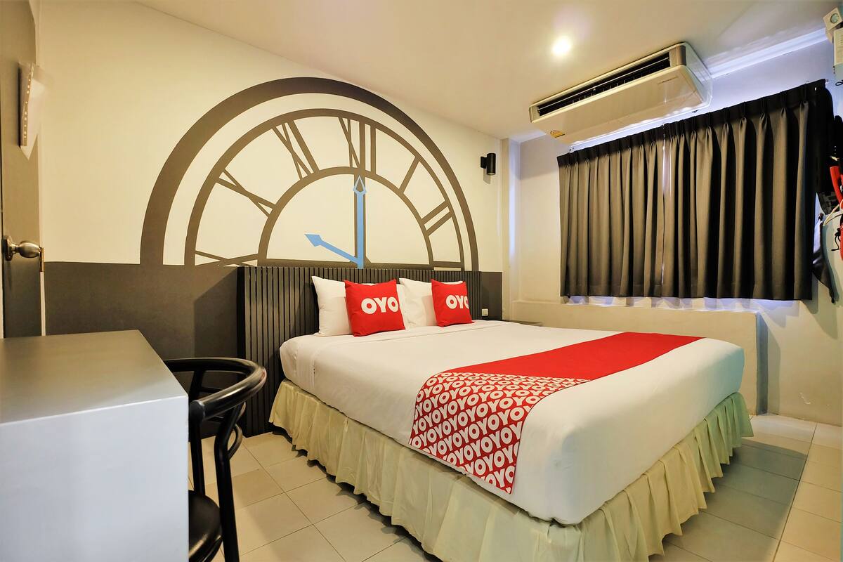 A hotel room with a bed, a desk, and a large wall clock.