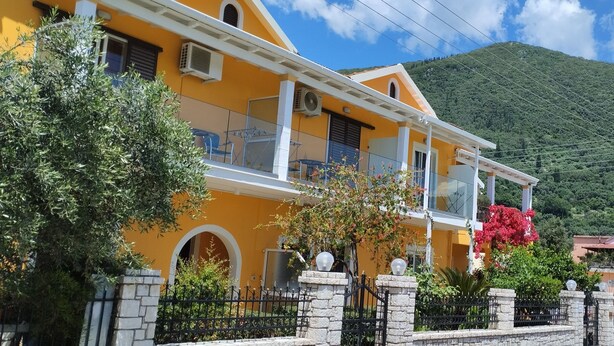 Un edificio giallo con un balcone, immerso nel verde con una montagna sullo sfondo.