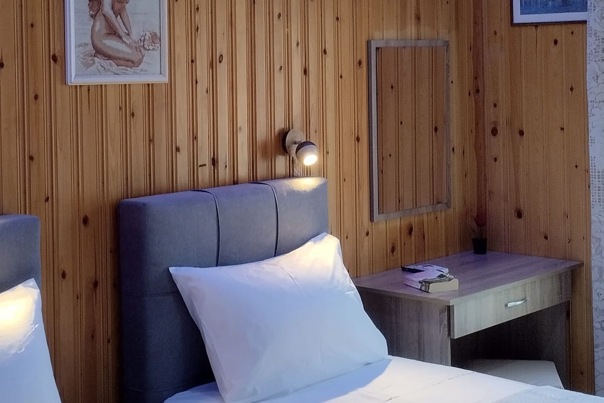 A hotel room with wooden panel walls, two beds with white linens, a bedside table with a lamp, and a framed picture on the wall.