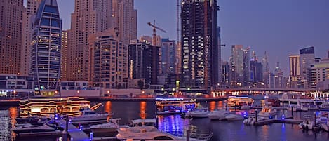 A marina with boats docked, a city skyline in the background, and a bridge connecting two areas.