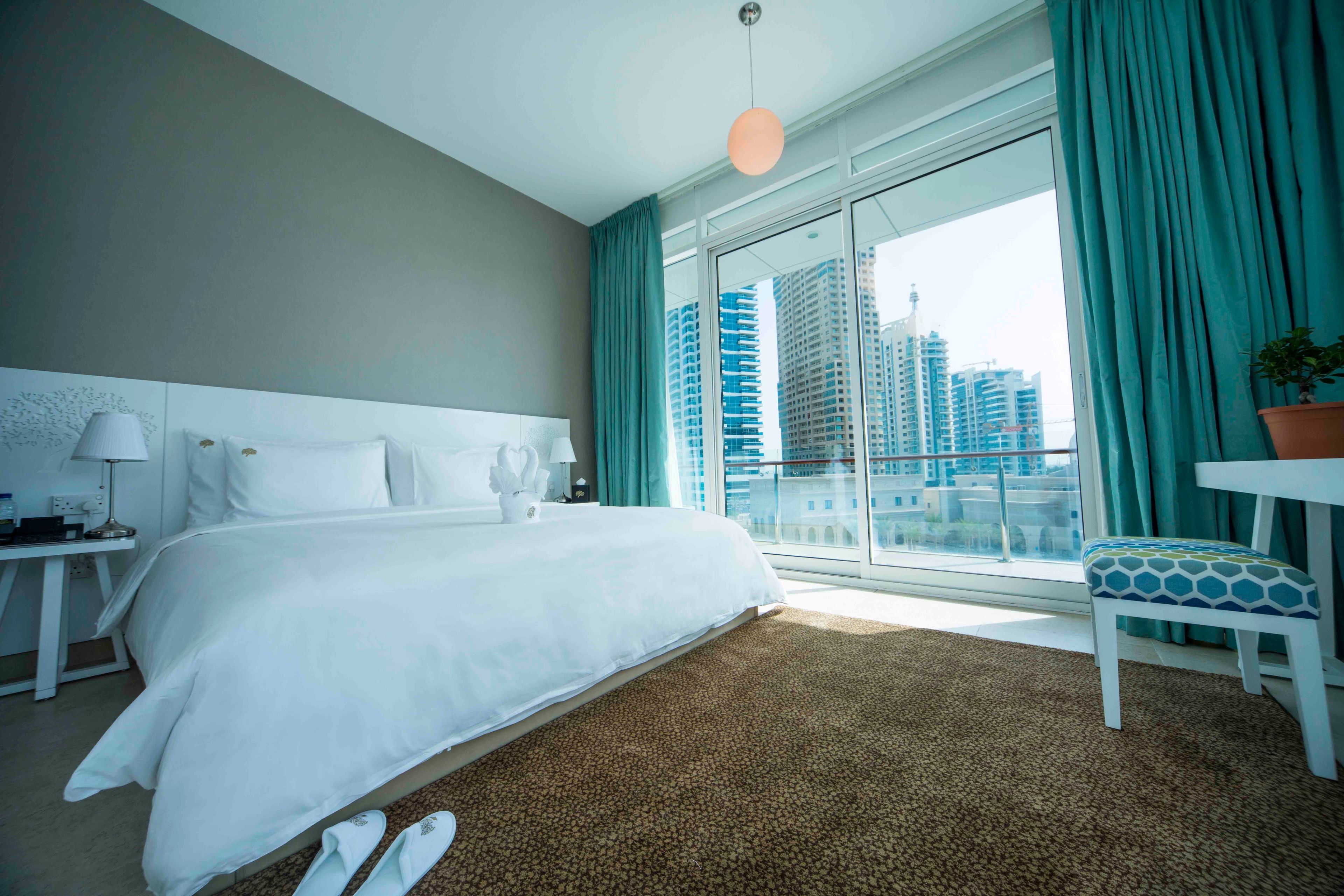 A hotel room with a large bed, a nightstand, a desk, a chair, and a view of tall buildings through the window.