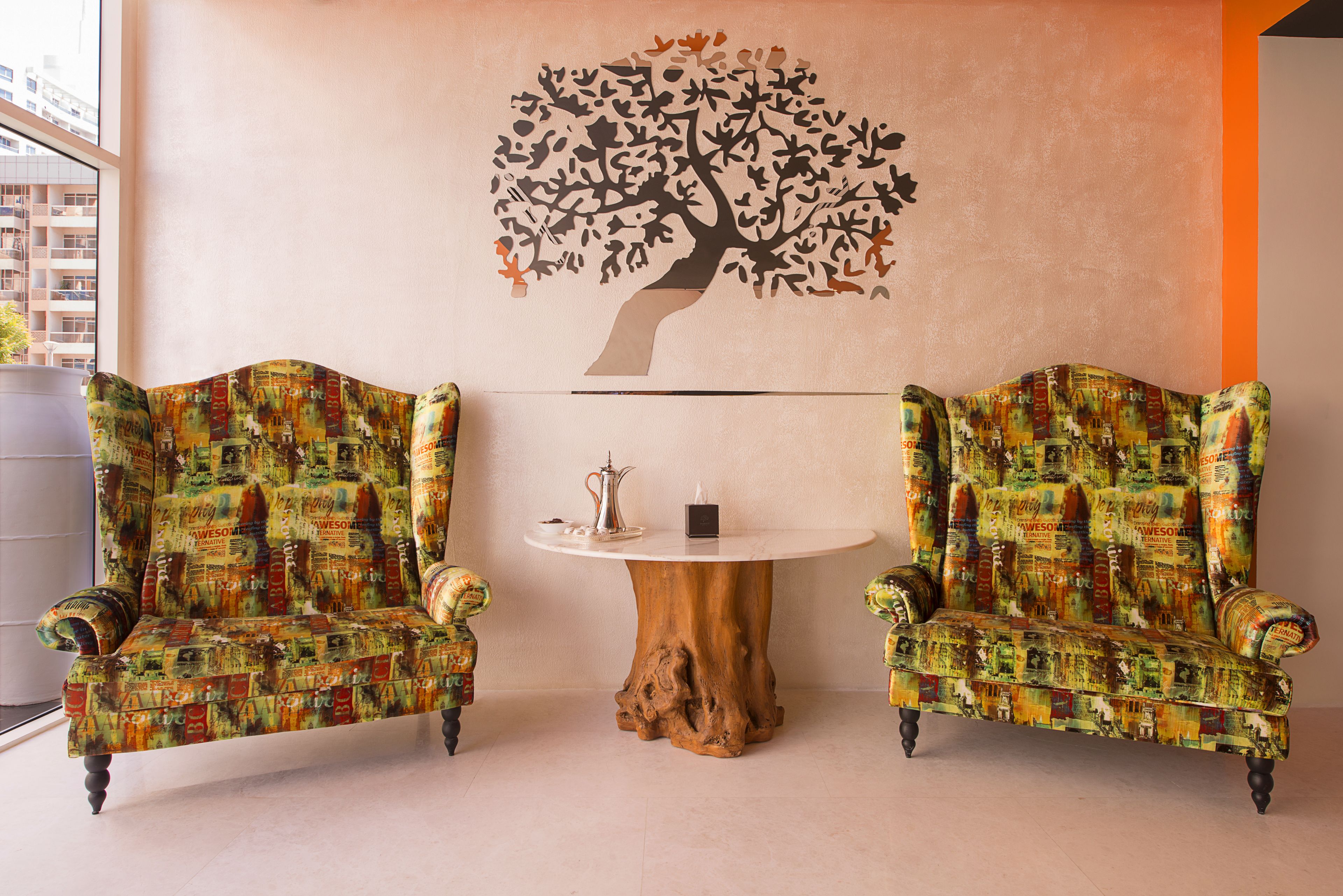 Dos sillones con tapicería estampada, una mesa redonda de madera con un pequeño adorno y un mural de un árbol en la pared.