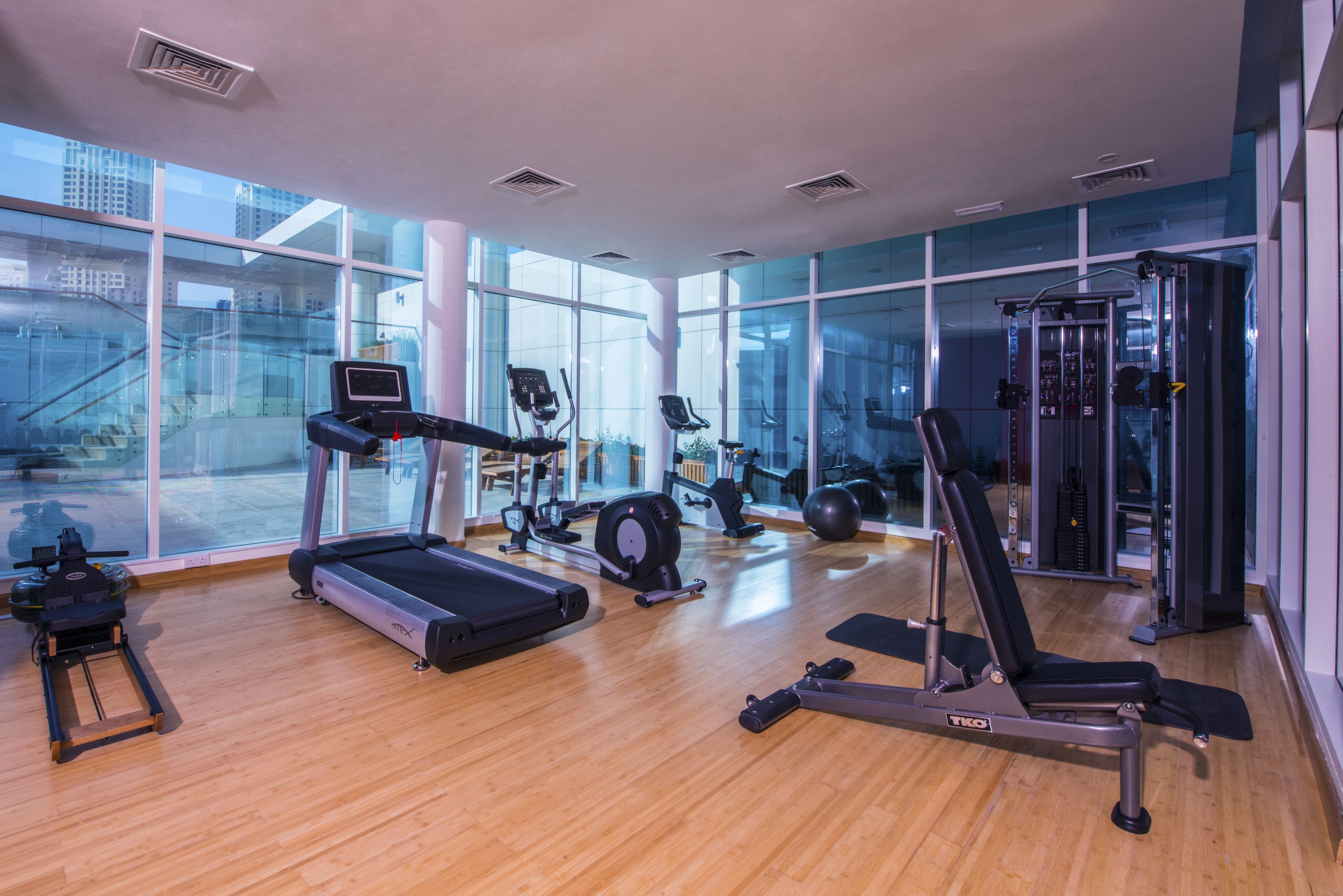 A modern gym with cardio equipment, weight machines, and a large window offering a city view.