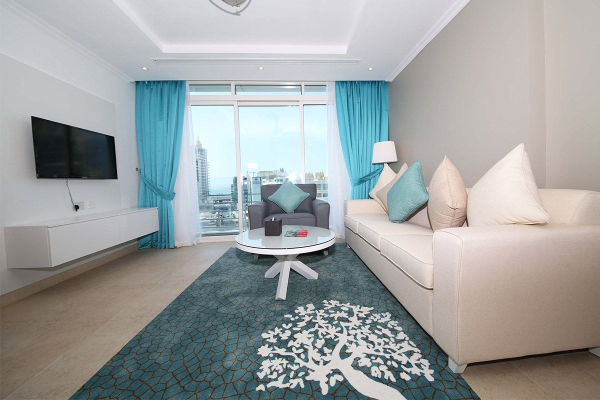 A modern hotel room with a large window, a flat-screen TV, a round coffee table, a sofa, and a patterned rug.